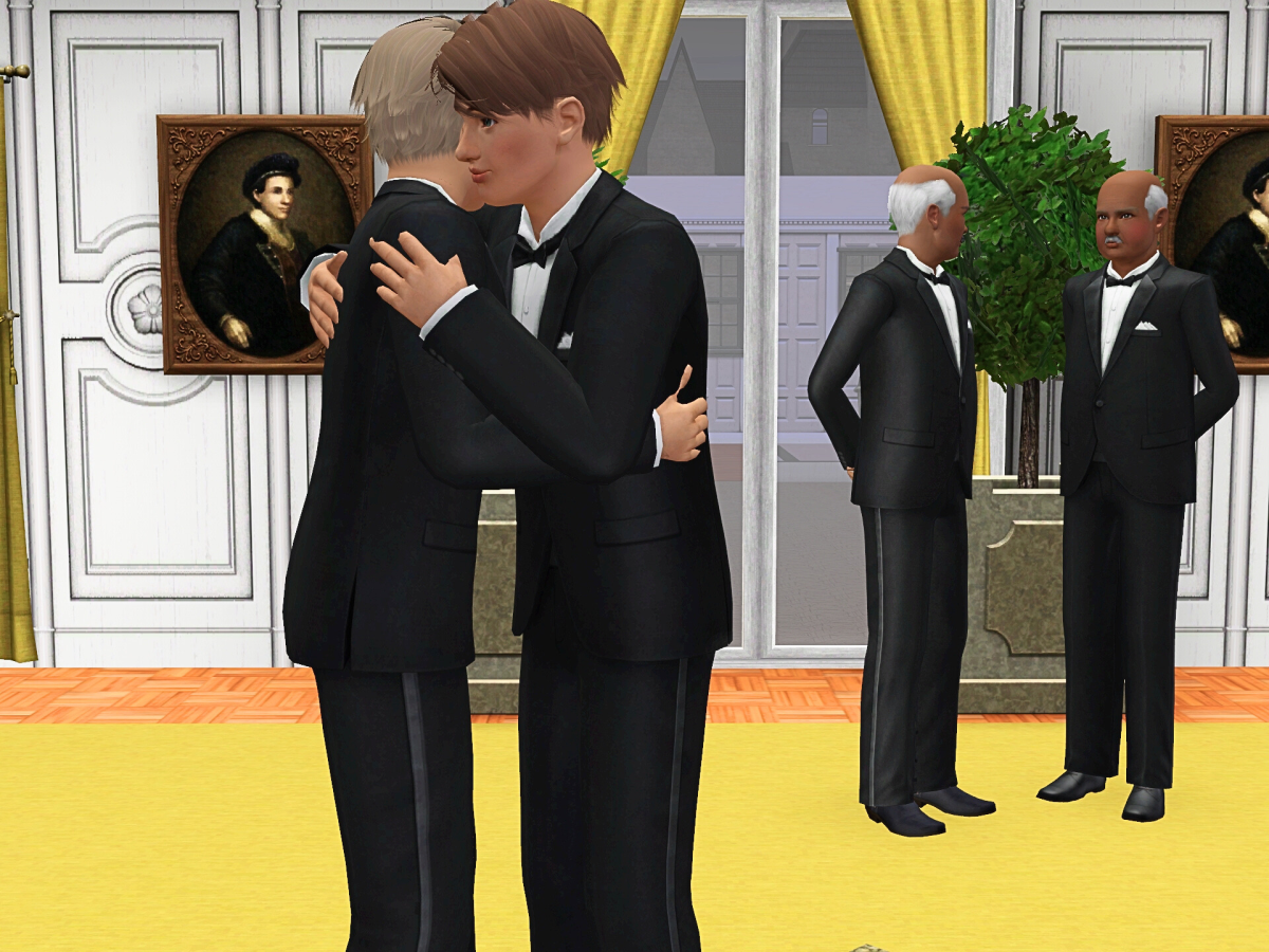 Two men in formal attire embrace in a warmly decorated room, with two other men standing nearby, all dressed in tuxedos. Artworks are displayed on the walls.