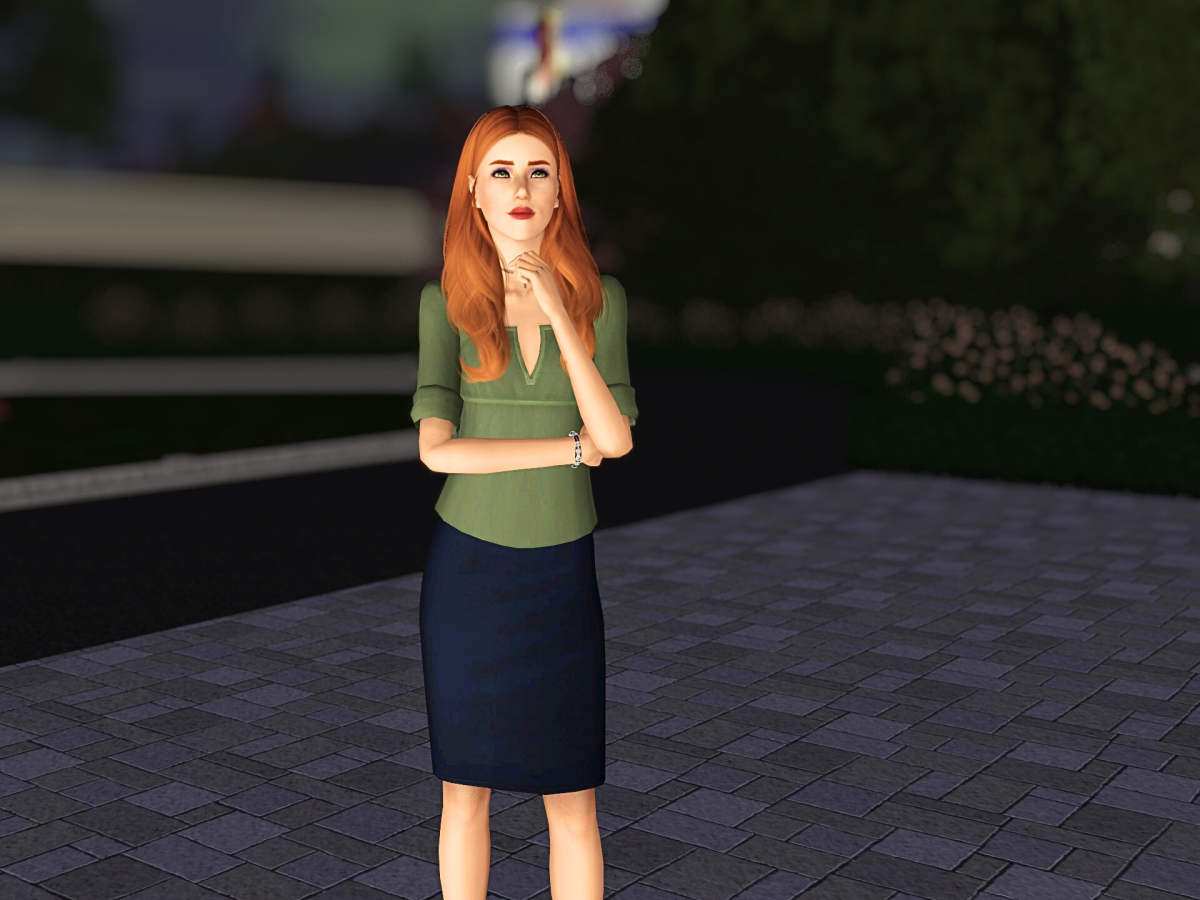 A woman with long red hair stands thoughtfully outdoors, wearing a green blouse and a dark skirt, on a paved surface with greenery in the background.