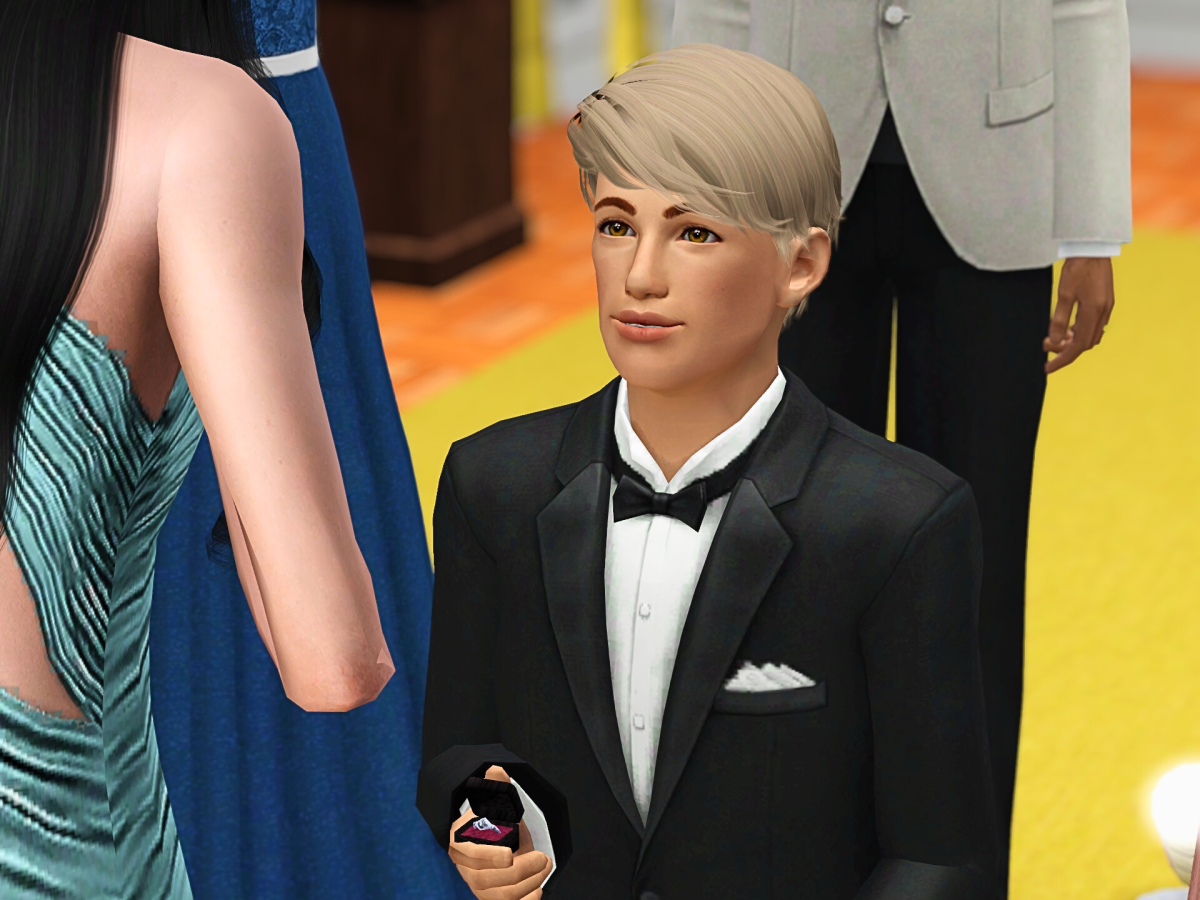 A young man in a tuxedo kneels, holding a ring in a box, gazing up at a woman in a teal dress. In the background, other guests are present at a festive event.