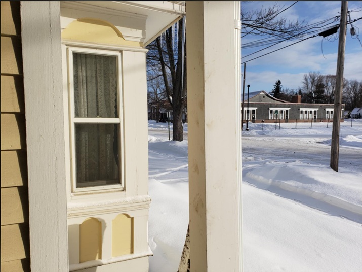 View from a house porch showing a window with lace curtains, snow-covered ground, and a distant building across the street.