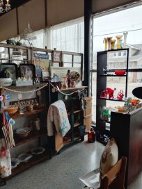 A cozy vintage shop interior featuring wooden shelving filled with various antique items, dishes, and decorative objects, alongside a display shelf with colorful collectibles and crafts.