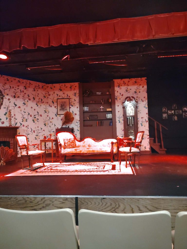 A theater stage set featuring a floral wallpaper backdrop, a vintage sofa, several wooden chairs, a coffee table with decorative items, an antique clock, and dim stage lighting.