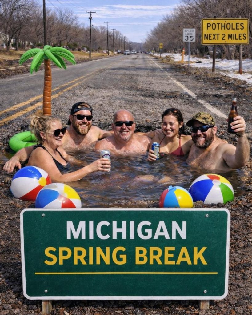 Four adults enjoying a hot tub in a roadside setting with a Michigan Spring Break sign, surrounded by beach balls and a palm tree, on a gravel road.