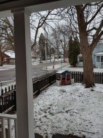 A snowy residential front yard with a view of a tree, a wooden fence, and houses across the street.