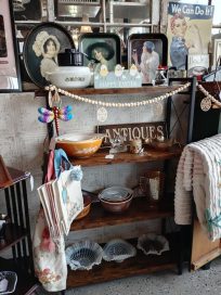 A wooden antique shelf displaying various vintage items, including bowls, decorative plates, and signs. The backdrop features framed pictures and decorative elements like a colorful butterfly ornament.