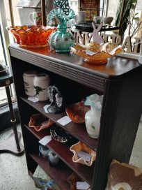 A wooden display shelf filled with various decorative items, including colored glass bowls, pottery, and unique trinkets.