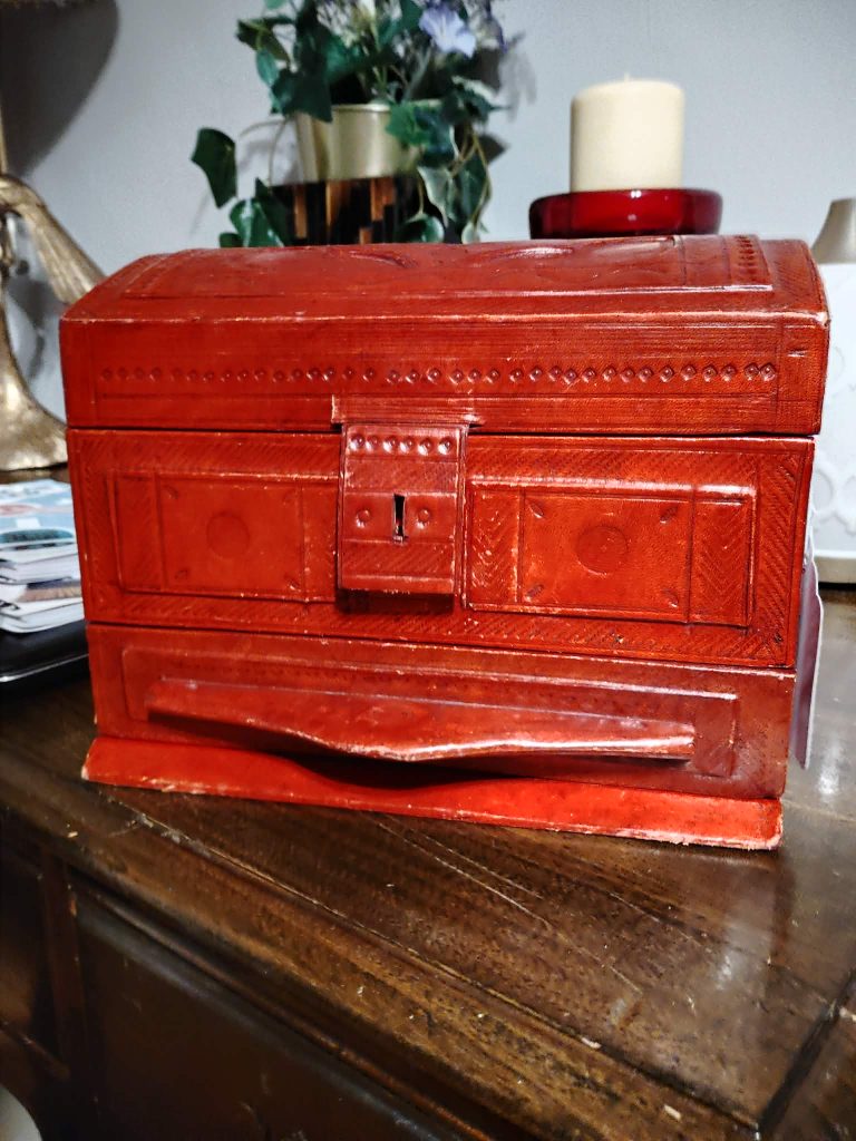 A decorative red wooden box with intricate carvings, featuring a small lock and a handle, placed on a wooden surface with a candle and plants in the background.