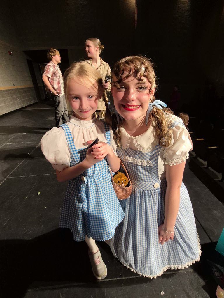 Two young girls dressed as characters from a musical, both wearing blue and white checkered dresses and smiling at the camera.