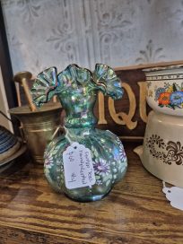 A vintage green glass vase with a ruffled top and floral patterns, displayed on a wooden surface among other antiques.