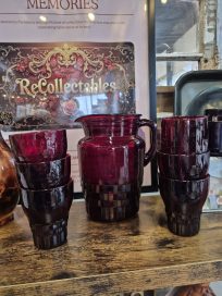 A collection of burgundy glassware consisting of a pitcher and several cups, displayed on a wooden surface with decorative items in the background.
