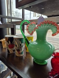 A display of decorative items featuring a green glass vase with a wavy rim and a handle, alongside cartoon character mugs with whimsical designs.