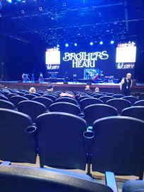 View of an empty concert hall with rows of seats facing a stage featuring the illuminated text 'BROTHERS HEART' and band equipment set up.