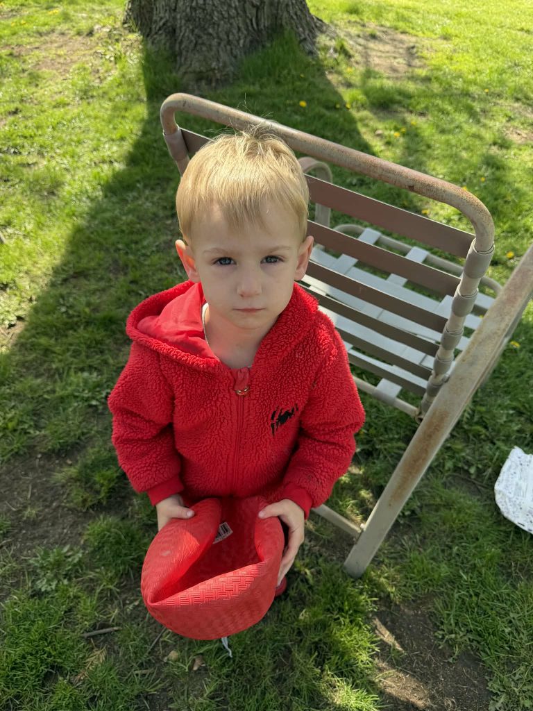 A young child in a red fleece jacket stands outdoors, holding a red hat, with a grassy background and a chair behind them.