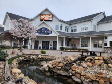 Exterior view of a restaurant with a sign and blooming cherry blossom tree in front, surrounded by a landscaped pond with rocks.