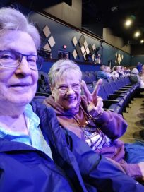Two older adults sitting in a theater with empty seats in the background, one making a peace sign and smiling.