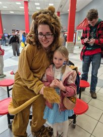 A person in a lion costume posing with a young girl dressed in a blue and white dress, in a vibrant indoor setting with red benches.