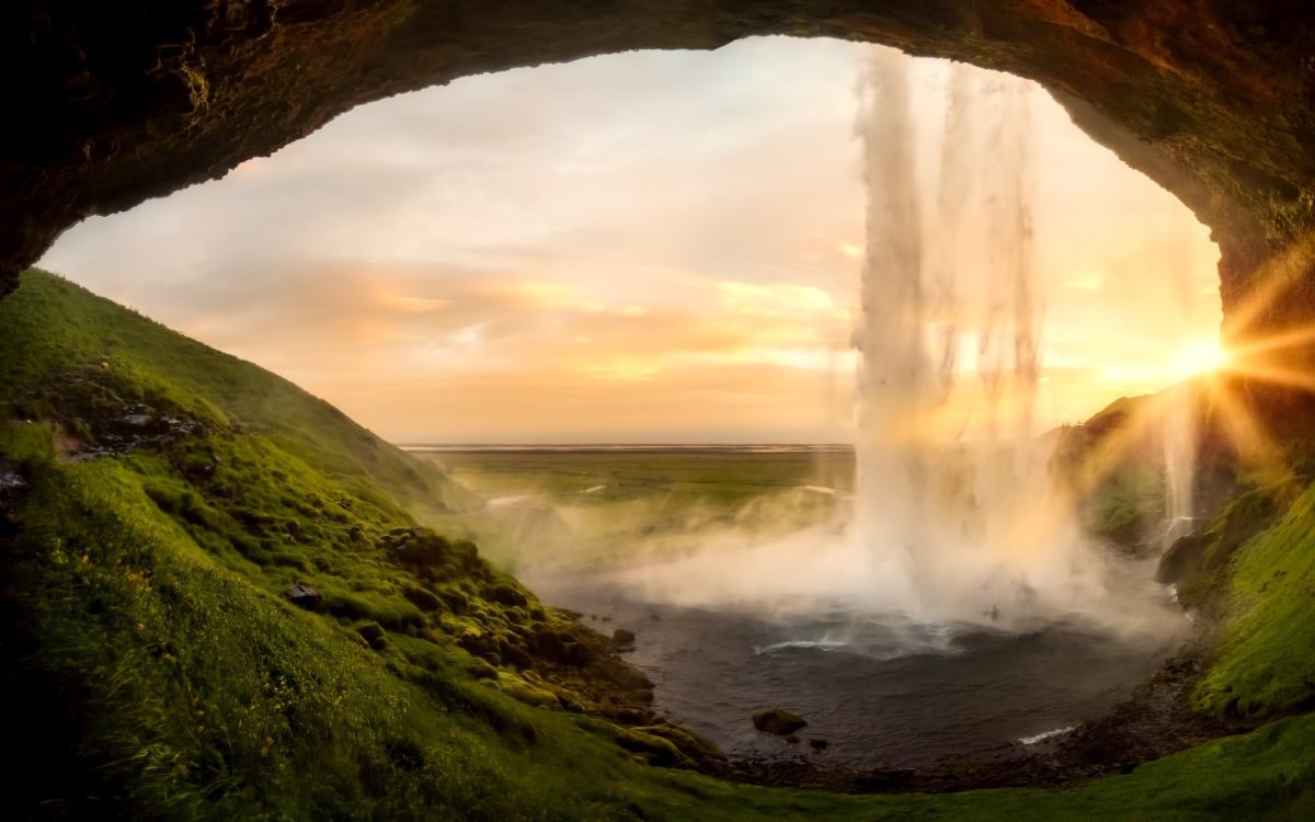 A scenic view from behind a waterfall, showcasing cascading water and a vibrant sunset in the background, surrounded by lush green hills.