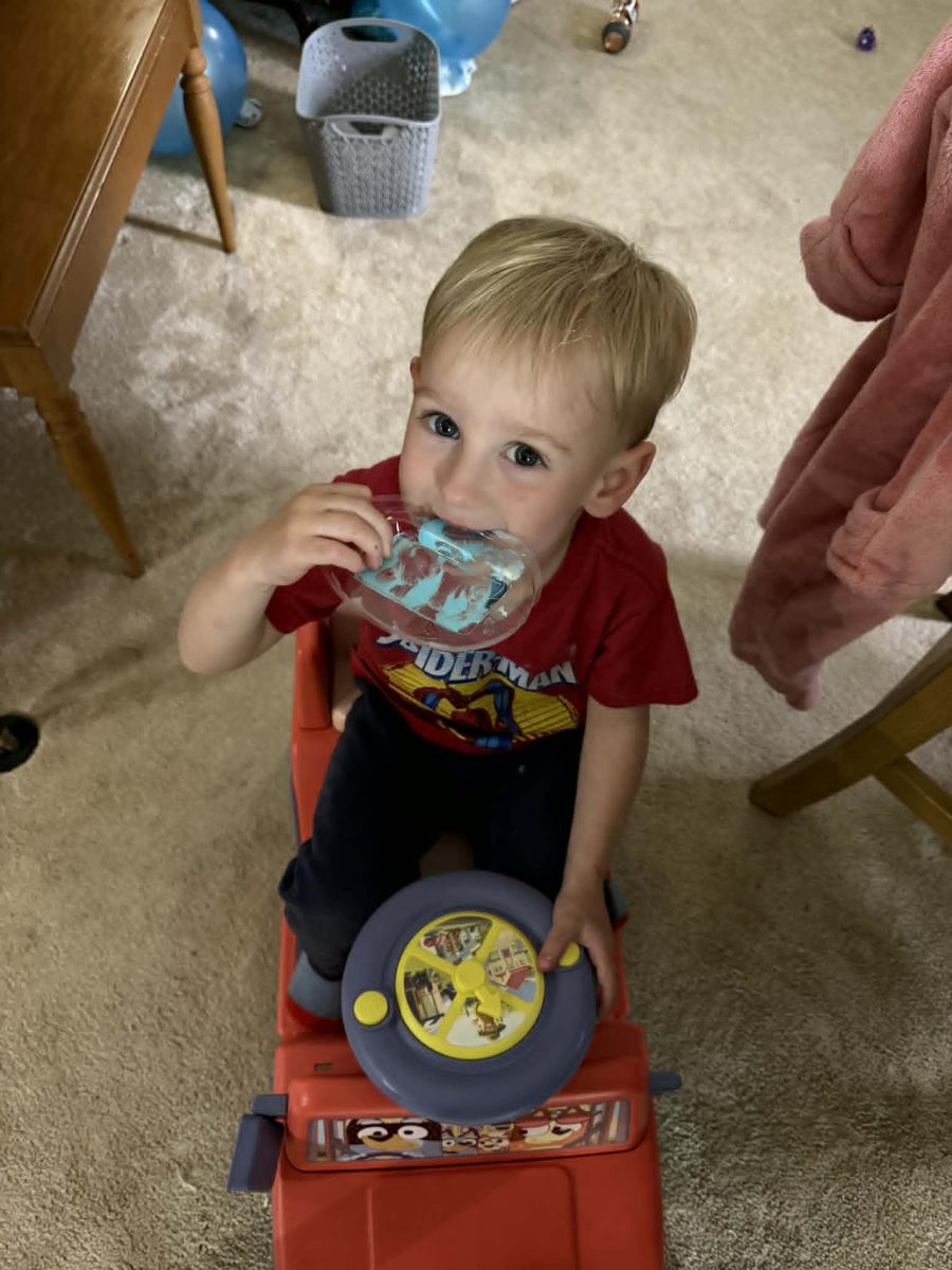 A young child sitting on a toy car, holding a colorful snack while wearing a red T-shirt.