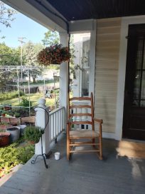 A wooden rocking chair on a porch, with a potted flower hanging nearby and a view of a garden with plants in pots in the background.