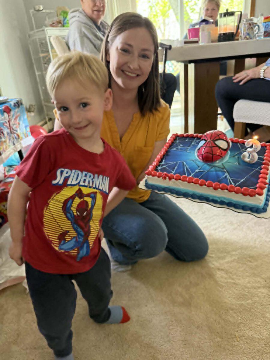 A young boy wearing a Spider-Man t-shirt poses with a smiling woman holding a decorated birthday cake featuring a Spider-Man design, in a festive indoor setting.