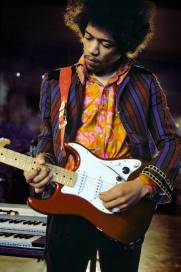 A male guitarist with an afro hairstyle playing an electric guitar while wearing a colorful patterned shirt and striped jacket.